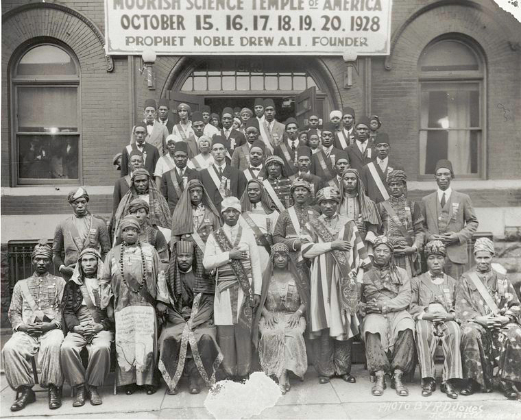 A group of Black men and women pose outside of the Moorish Science Temple of America and under a banner. The banner states the name of the temple, and the dates October 15-20, 1928. Below that is written "Prophet Noble Ali. Founder."